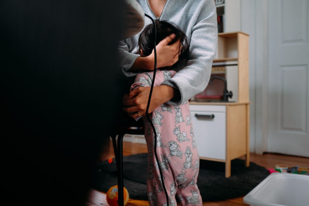 An image of a small child with black hair hugging their parent. The child is wearing light pink pajamas with grey cartoon bunnies on them.